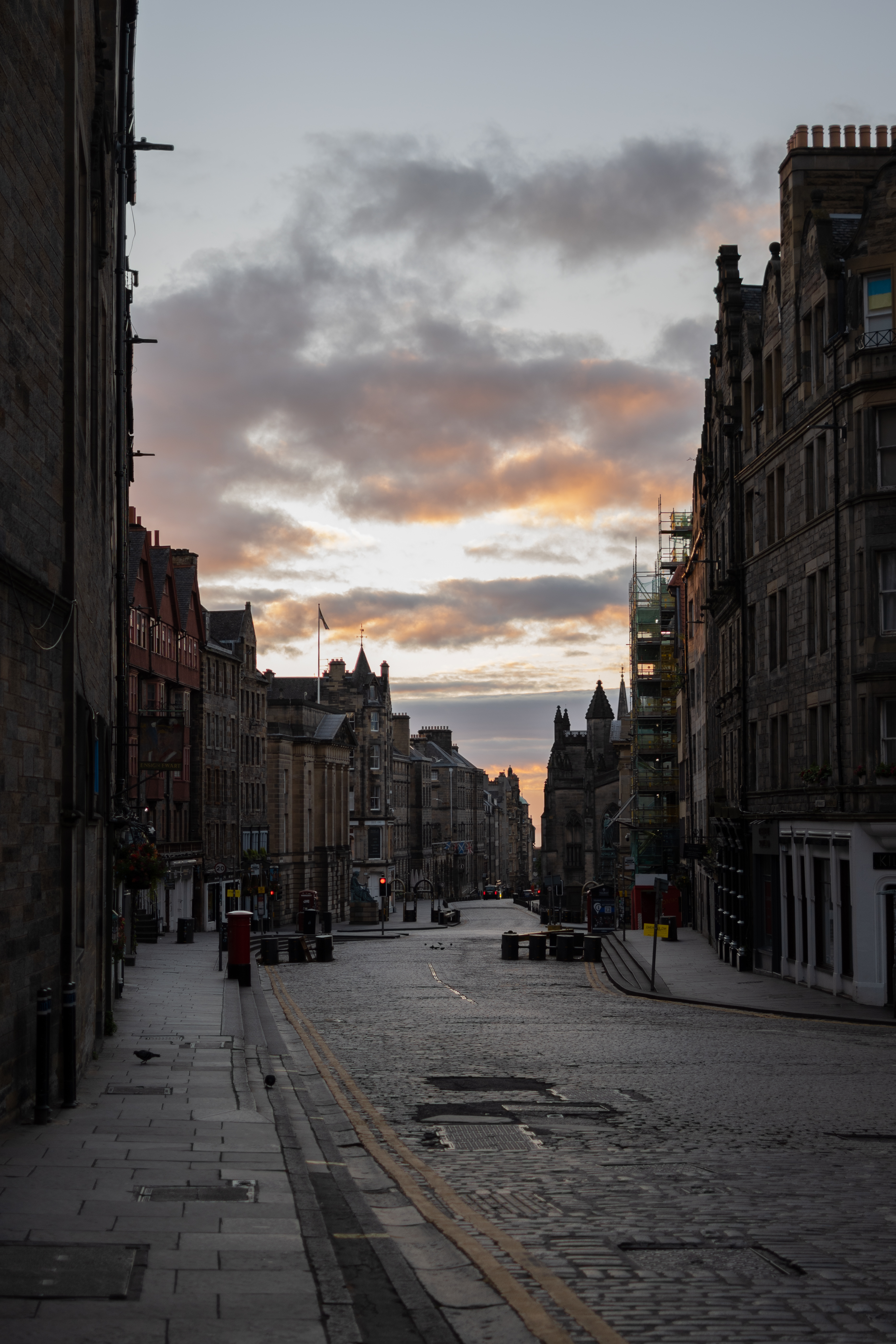 A portrait aspect scene of a curving street. The road is cobbled with double yellow lines along it and a pavement on both sides. The road is flanked by tall old buildings of different heights. Many have towers and chimneys on top of them and pointed rooves. Towards the middle of the image along the road there is a red post box on the left and traffic lights showing red lights. The sky has bands of clouds which are tinged with a peachy gold colour from the rising sun. The rest of the clouds are grey and sky behind them is an extremely pale blue almost white. 